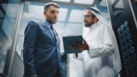Téléchargez les photos : Homme d'affaires européen Parler avec un partenaire d'investissement arabe tout en chevauchant ascenseur en verre au bureau dans un centre d'affaires moderne. Société internationale - en image libre de droit