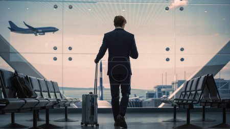 Téléchargez les photos : Terminal de l'aéroport : Homme d'affaires avec valise roulante Promenades, Utilise l'application Smartphone pour les affaires électroniques. Silhouette de l'homme voyageur attend pour le vol - en image libre de droit