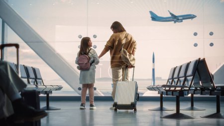 Foto de Terminal del aeropuerto: Hermosa madre y linda hijita esperan su vuelo de vacaciones, mirando por la ventana para llegar y salir de los aviones - Imagen libre de derechos