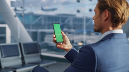 Foto de Terminal del aeropuerto: Empresario que trabaja en Green Chroma Key Screen Mobile Smartphone, Espera un vuelo en avión. Empresario hace trabajo remoto en línea en - Imagen libre de derechos