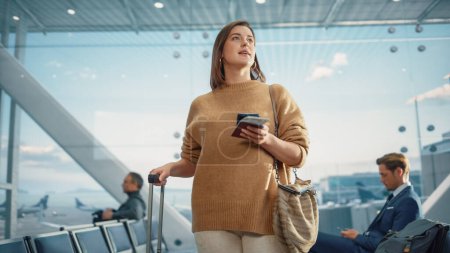 Foto de Terminal del aeropuerto: Mujer caucásica viajando feliz que espera en las puertas de vuelo para el embarque del avión, utiliza el teléfono inteligente móvil, chequeando el destino del viaje en - Imagen libre de derechos