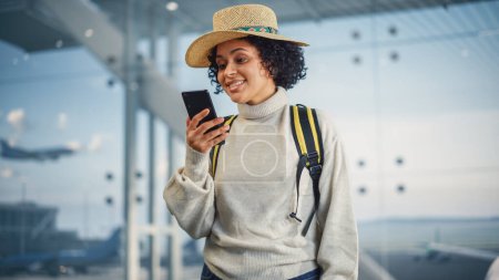 Foto de Terminal del aeropuerto: Mujer negra viajera feliz mira alrededor buscando puertas de vuelo y avión, utiliza Smartphone, Comprobación de destino de viaje en Internet - Imagen libre de derechos
