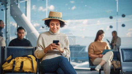 Foto de Terminal del aeropuerto: Mujer negra feliz espera para el vuelo, Utiliza Smartphone, Navegar por Internet, Publicar en las redes sociales, Compras en línea. Viajar por África - Imagen libre de derechos