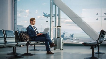 Téléchargez les photos : Terminal de l'aéroport moderne : bel homme d'affaires travaillant sur ordinateur portable en attendant son vol. Homme assis dans un salon d'embarquement de Big Airline - en image libre de droit