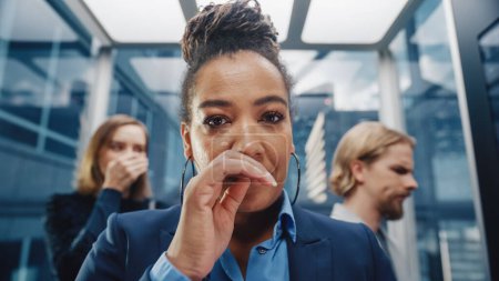 Photo for Close Up Portrait of a Middle Aged Woman Sneezes in a Crowded Glass Elevator in a Modern Office Building. Businesswoman Covers Her Face, but Other - Royalty Free Image