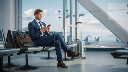 Téléchargez les photos : Terminal de l'aéroport : Homme d'affaires utilise Smartphone, En attente d'un vol, Faire des affaires en ligne, Naviguer sur Internet avec une application. Entrepreneur itinérant - en image libre de droit