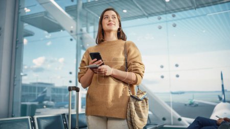 Téléchargez les photos : Terminal de l'aéroport : Happy Traveling Femme caucasienne en attente aux portes de l'avion pour l'embarquement, Utilise Smartphone mobile, Vérification de la destination du voyage sur - en image libre de droit