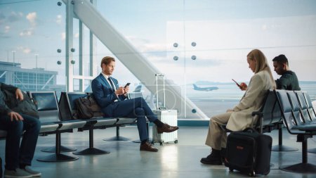Téléchargez les photos : Terminal de l'aéroport occupé : bel homme d'affaires utilise Smartphone en attendant son vol. Personnes assises dans un salon d'embarquement de Big Airline Hub avec - en image libre de droit
