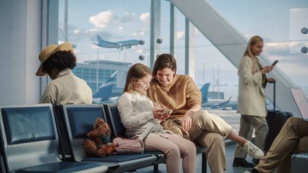 Photo for Busy Airport Airplane Terminal: Happy Beautiful Mother and Cute Little Daughter Wait for their Vacation Flight, Use Mobile Smartphone for Fun. Diverse - Royalty Free Image