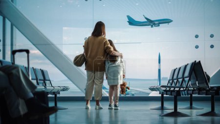Téléchargez les photos : Terminal de l'aéroport : Belle mère et jolie petite fille attendent leur vol de vacances, regardant par la fenêtre pour l'arrivée et le départ des avions - en image libre de droit
