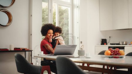 Foto de Hermosa mamá joven sosteniendo a su precioso hijo bebé, utilizando la computadora portátil en la sala de estar en el apartamento. Madre está trabajando desde casa, Compras en línea - Imagen libre de derechos