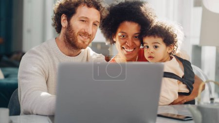 Foto de El padre, la madre y el hijo joven usan la computadora portátil en Home Apartment. Familia de raza mixta feliz con dibujos animados del reloj del niño cariñoso, tienda productos del bebé - Imagen libre de derechos