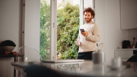 Photo for Handsome Adult Man with Ginger Curly Hair Using Smartphone, Standing in Living Room at Home. Male Enjoys a Cup of Espresso, Checking Social Media - Royalty Free Image