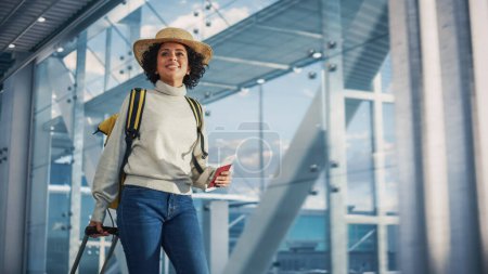 Téléchargez les photos : Terminal de l'aéroport : Belle femme noire souriante détient un billet, se promène dans le carrefour de la compagnie aérienne jusqu'aux portes où l'avion attend. Heureux Afro-américain - en image libre de droit