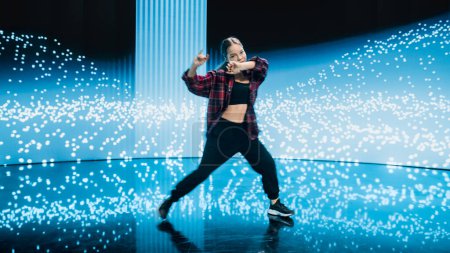 Téléchargez les photos : Jeune danseuse professionnelle élégante exécutant une routine de danse hip-hop devant un grand écran mural mené avec animation VFX lors d'un virtuel - en image libre de droit