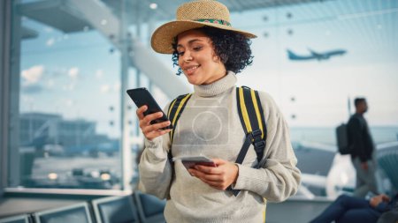 Foto de Terminal del aeropuerto: Mujer negra viajando feliz que espera en las puertas de vuelo para el embarque del avión, utiliza el teléfono inteligente móvil, chequeando el destino del viaje en - Imagen libre de derechos