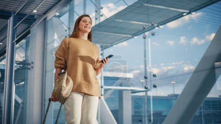 Photo for Airport Terminal: Beautiful Smiling Woman Holds Smartphone, Walks in Airline Hub to the Gates Where Airplane Waits Her. Happy Caucasian Female is - Royalty Free Image