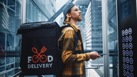 Photo for Young Food Delivery Person Riding Glass Elevator in Modern Office Building. Restaurant Delivery Courier Using Smartphone to Check Order. Handsome - Royalty Free Image