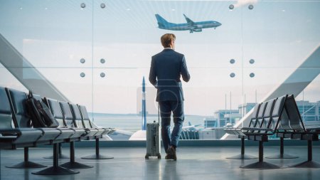 Photo for Airport Terminal: Businessman with Rolling Suitcase Walks, Uses Smartphone App for e-Business. Back View of Traveling Man Waiting for a Flight in - Royalty Free Image