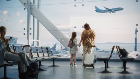 Foto de Terminal del aeropuerto: Hermosa madre y linda hijita esperan su vuelo de vacaciones, mirando por la ventana para llegar y salir de los aviones - Imagen libre de derechos