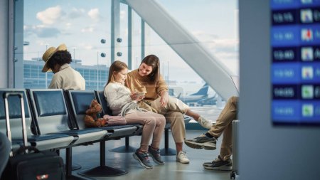 Foto de Terminal de aviones del aeropuerto ocupado: Happy Beautiful Mother y Cute Little Daughter Espere a que su vuelo de vacaciones, utilice el teléfono inteligente móvil para divertirse. Diversos - Imagen libre de derechos