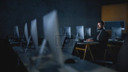Téléchargez les photos : Étudiant masculin concentré assis seul dans la salle informatique du collège, soirée de travail sur le projet informatique. Jeune érudit Étude IT sur - en image libre de droit