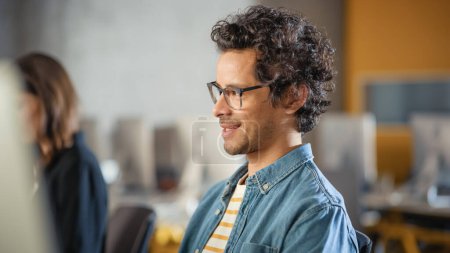 Retrato de un apuesto estudiante masculino inteligente, estudiando en la universidad, sonriendo cariñosamente. Lleva gafas y tiene el pelo rizado oscuro. Trabaja en la computadora en
