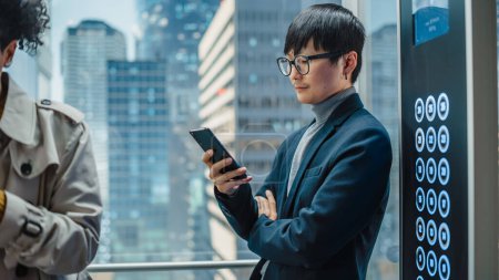 Photo for Successful Stylish Japanese Businessman Riding Glass Elevator to Office in Modern Business Center. Handsome Man Smile while Using Smartphone, Write - Royalty Free Image