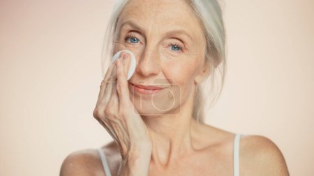 Foto de Medium Potrait: Beautiful Senior Woman utiliza almohadilla de algodón para limpiar su perfecta cara de cosméticos. Una anciana sonriente con piel suave. Viejo agraciado - Imagen libre de derechos