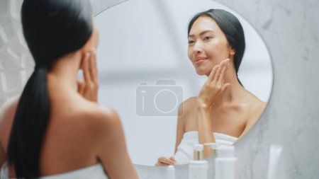 Portrait der schönen asiatischen Frau sanft auftragen Gesichtscreme Maske mit sinnlichen Berührungen, Blick in Badezimmerspiegel. Weiblich macht ihre Haut weich mit