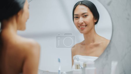 Téléchargez les photos : Portrait of Beautiful Asian Woman Gently Looking in Bathroom Mirror. Young Female with Skin Soft, Smooth, Wrinkle Free with Natural anti-aging - en image libre de droit