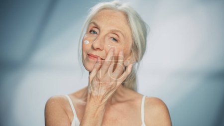 Foto de Retrato de una hermosa mujer mayor aplicando suavemente crema facial. La anciana Lady hace que su piel sea suave, lisa, sin arrugas con antienvejecimiento natural - Imagen libre de derechos