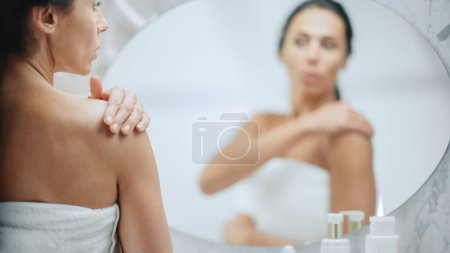 Schöne kaukasische Frau trägt Körpercreme auf ihre perfekte Schulter auf und macht die Haut weich, sieht im Badezimmerspiegel aus. Glücklich weiblich genießen sie schönheit