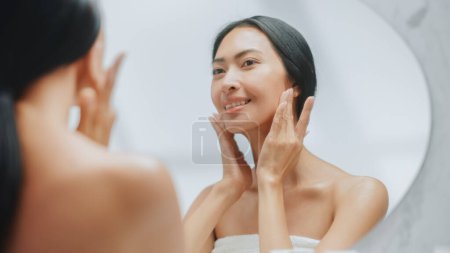 Porträt der schönen Asiatin, die sanft Gesichtscreme im Badezimmerspiegel aufträgt. Junge erwachsene Frau macht ihre Haut weich, glatt und natürlich
