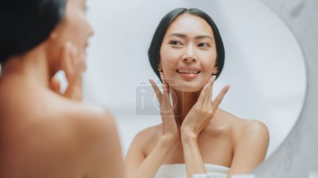 Porträt der schönen Asiatin, die sanft Gesichtscreme im Badezimmerspiegel aufträgt. Junge Frau macht ihre Haut weich, glatt, faltenfrei mit