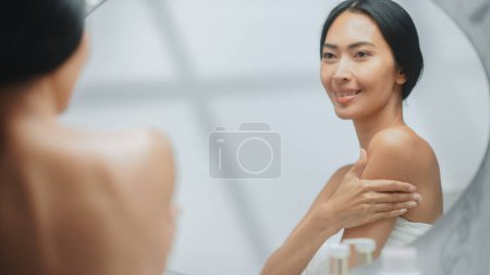 Schöne Asiatin berührt ihre perfekte weiche Schulter, ihren Hals und lächelt sinnlich in den Spiegel. Glückliche Weibchen genießen ihre Schönheit. Wellness Natur