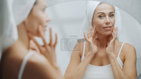 Foto de Beautiful Caucasian Woman Touches Her Perfect Soft Shoulder, Neck, Sensually Looks in Bathroom Mirror. Middle Aged Female Enjoying Her Natural Beauty - Imagen libre de derechos