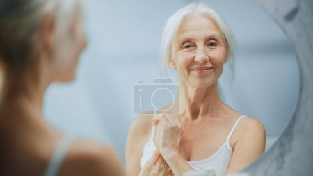 Foto de Retrato de hermosa mujer mayor rutina de la mañana, Mirando en espejo suavemente aplicación de crema facial. anciana dama hace su piel suave, suave, arrugas - Imagen libre de derechos