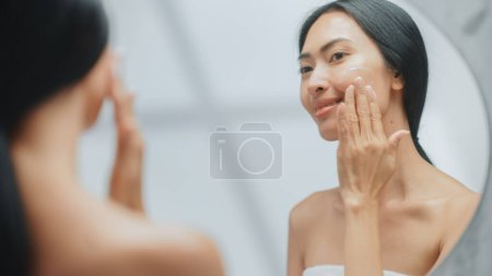 Portrait der schönen asiatischen Frau sanft auftragen Gesichtscreme Maske mit sinnlichen Berührungen, Blick in Badezimmerspiegel. Weiblich macht ihre Haut weich mit