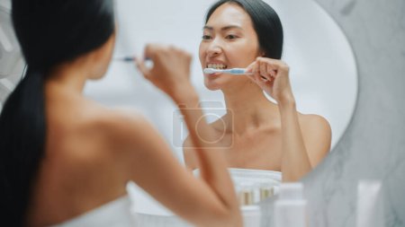 Téléchargez les photos : Portrait de belle femme asiatique utilise brosse à dents pour nettoyer ses dents blanches naturelles, sourire dans miroir de salle de bains en regardant la caméra. Fille heureuse faisant - en image libre de droit