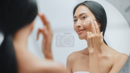 Portrait der schönen asiatischen Frau sanft auftragen Gesichtscreme Maske mit sinnlichen Berührungen, Blick in Badezimmerspiegel. Junge Frau macht ihre Haut weich