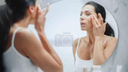 Porträt der schönen kaukasischen Frau, die sanft Gesichtscreme mit sinnlichen Gesten aufträgt und in den Badezimmerspiegel blickt. Weibliches Mittelalter macht Haut
