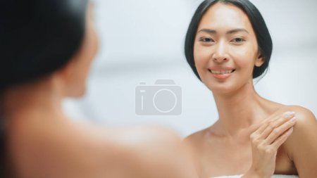 Porträt der schönen Asiatin, die sanft Körpercreme mit sinnlichen Berührungen aufträgt und in den Badezimmerspiegel blickt. Junge Frau macht die Haut weich mit