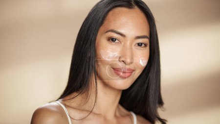 Photo for Female Beauty Portrait. Beautiful Asian Happy Brunette Woman Posing with Applied Face Cream. Wellness and Skincare Concept on Isolated Background. - Royalty Free Image