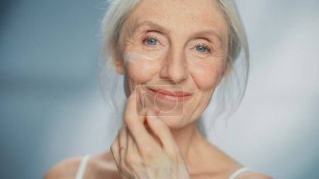Porträt der schönen Seniorin, die sanft Gesichtscreme aufträgt. Ältere Dame macht ihre Haut weich, glatt, faltenfrei mit natürlichem Anti-Aging