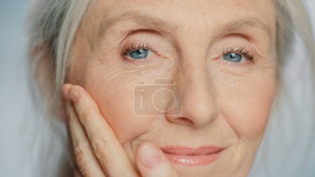 Téléchargez les photos : Gros plan Portrait d'une belle femme âgée qui applique doucement de la crème pour le visage. Dame âgée rend sa peau douce, lisse, sans rides avec anti-âge naturel - en image libre de droit