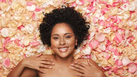 Foto de Retrato de belleza femenina acostado en cama de flores. Hermosa mujer afroamericana con cabello afro, natural, limpia, piel sana posando y sonriendo. Bienestar - Imagen libre de derechos