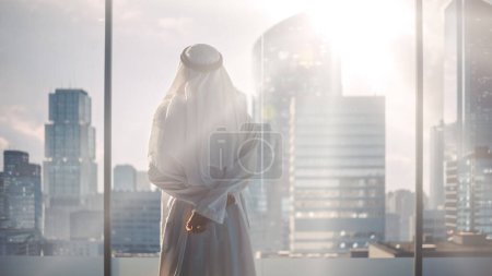 Téléchargez les photos : Homme d'affaires musulman prospère en tenue blanche traditionnelle debout dans son bureau moderne regardant par la fenêtre sur la grande ville avec des gratte-ciels - en image libre de droit