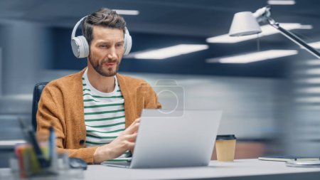 Photo for Modern Office: Handsome Businessman Sitting at His Desk Working on a Laptop Computer. Man Wearing Headphones, Listens to Music, Podcast. Motion Blur - Royalty Free Image