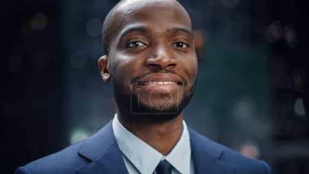 Téléchargez les photos : Portrait d'un homme d'affaires noir réfléchi portant une combinaison, debout dans la rue du quartier des affaires de la grande ville, regardant la caméra et souriant. Succès - en image libre de droit